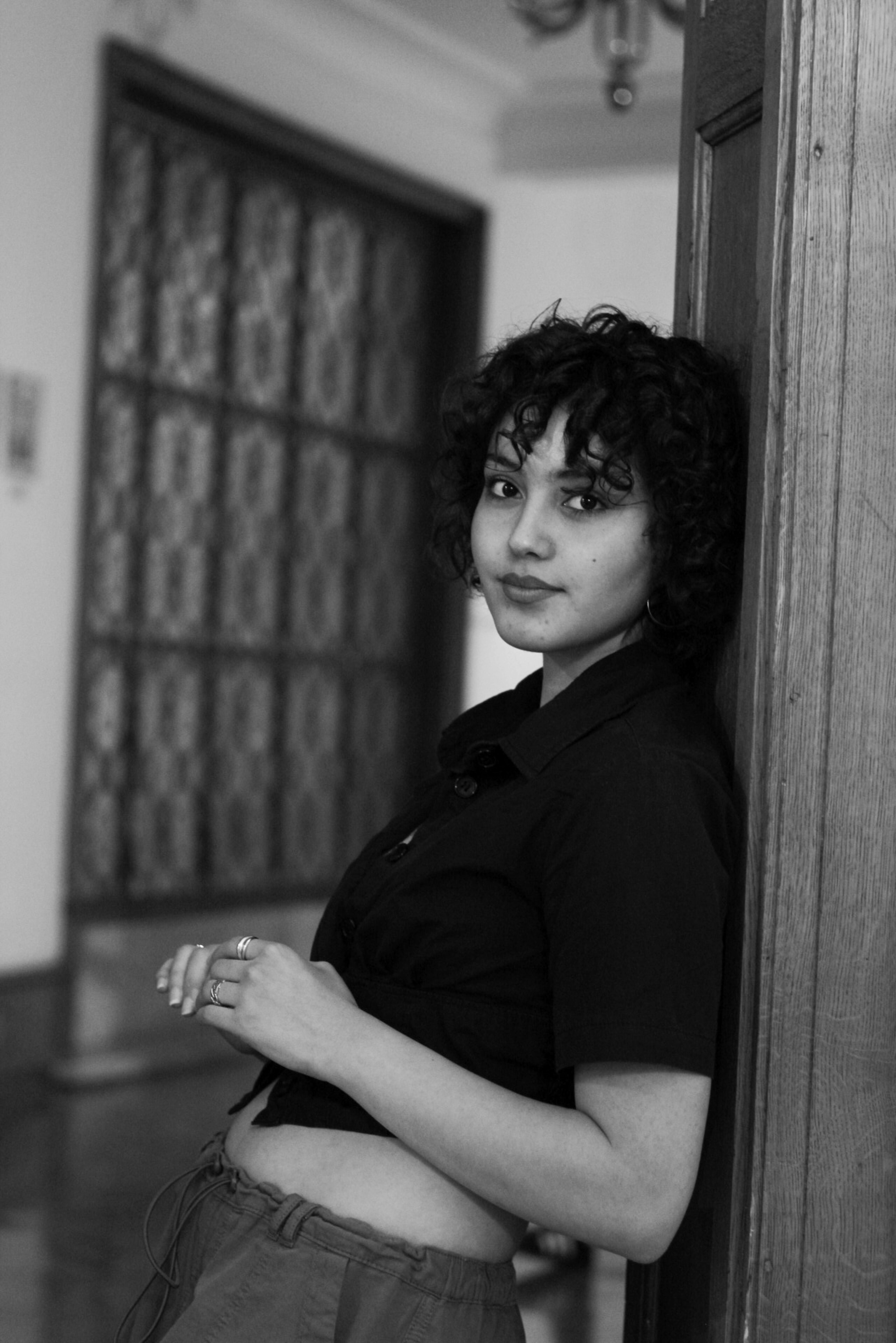 A young woman with curly hair smiling while leaning in a doorframe, captured in a monochrome setting.