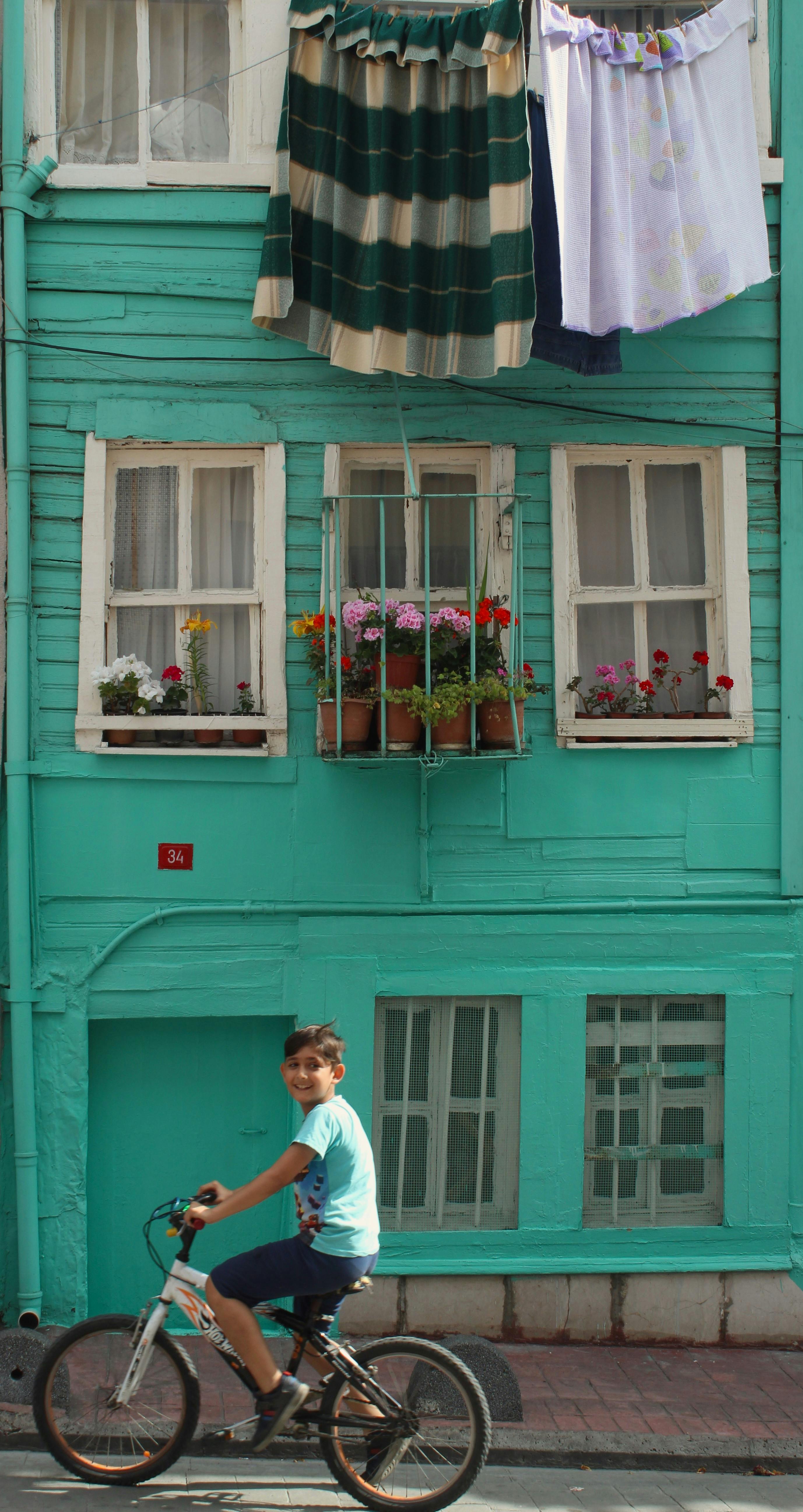 A young boy rides a bicycle past a vibrant teal building with hanging laundry and potted flowers.