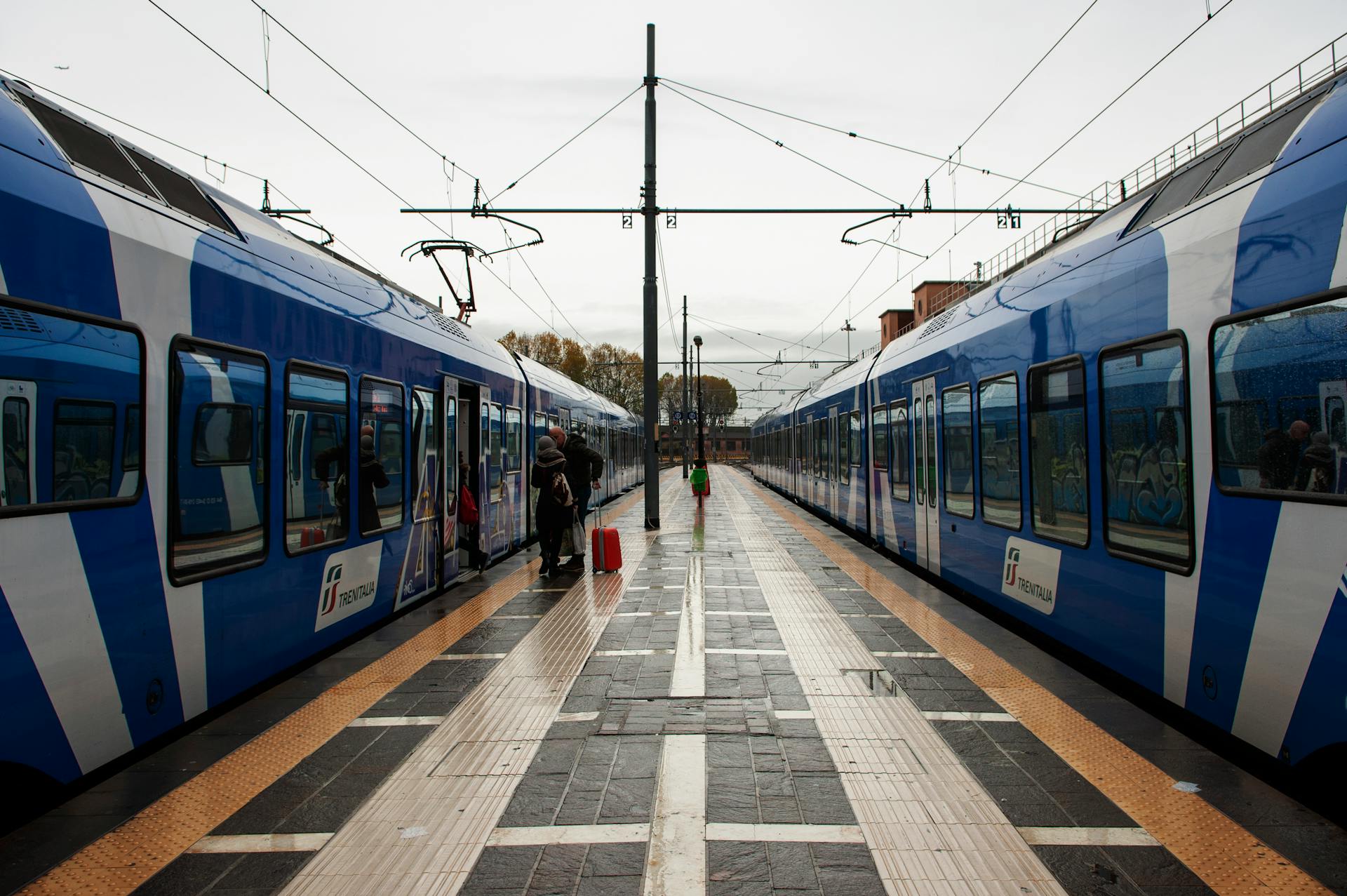 A symmetrical view of a railway platform with Trenitalia trains at a station with passengers boarding.