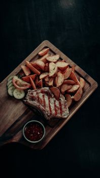 Grilled steak with potato wedges, cucumbers, and sauce on a wooden board.