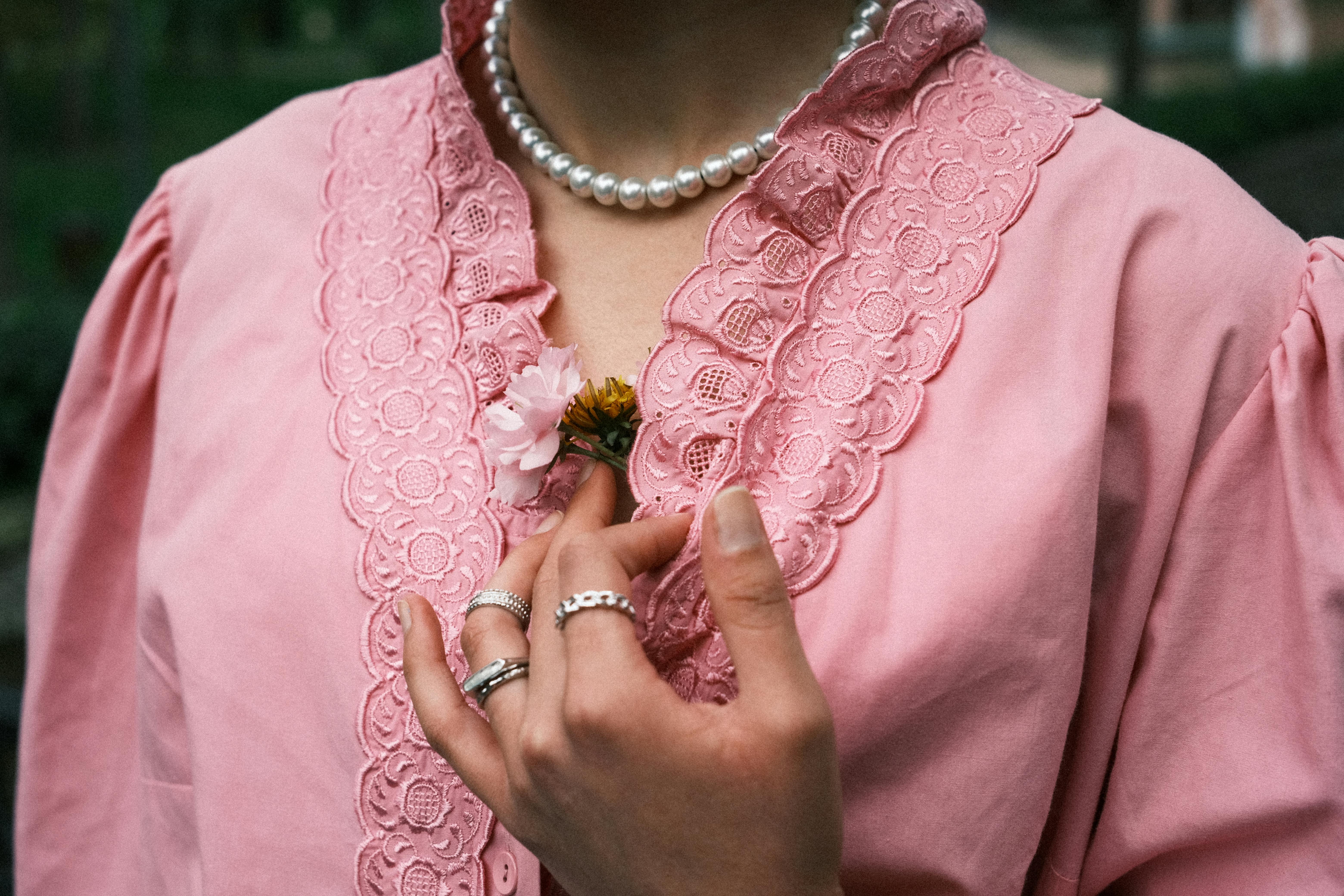 A woman's hand holds a flower against a pink lace blouse, featuring elegant pink attire and pearl necklace.