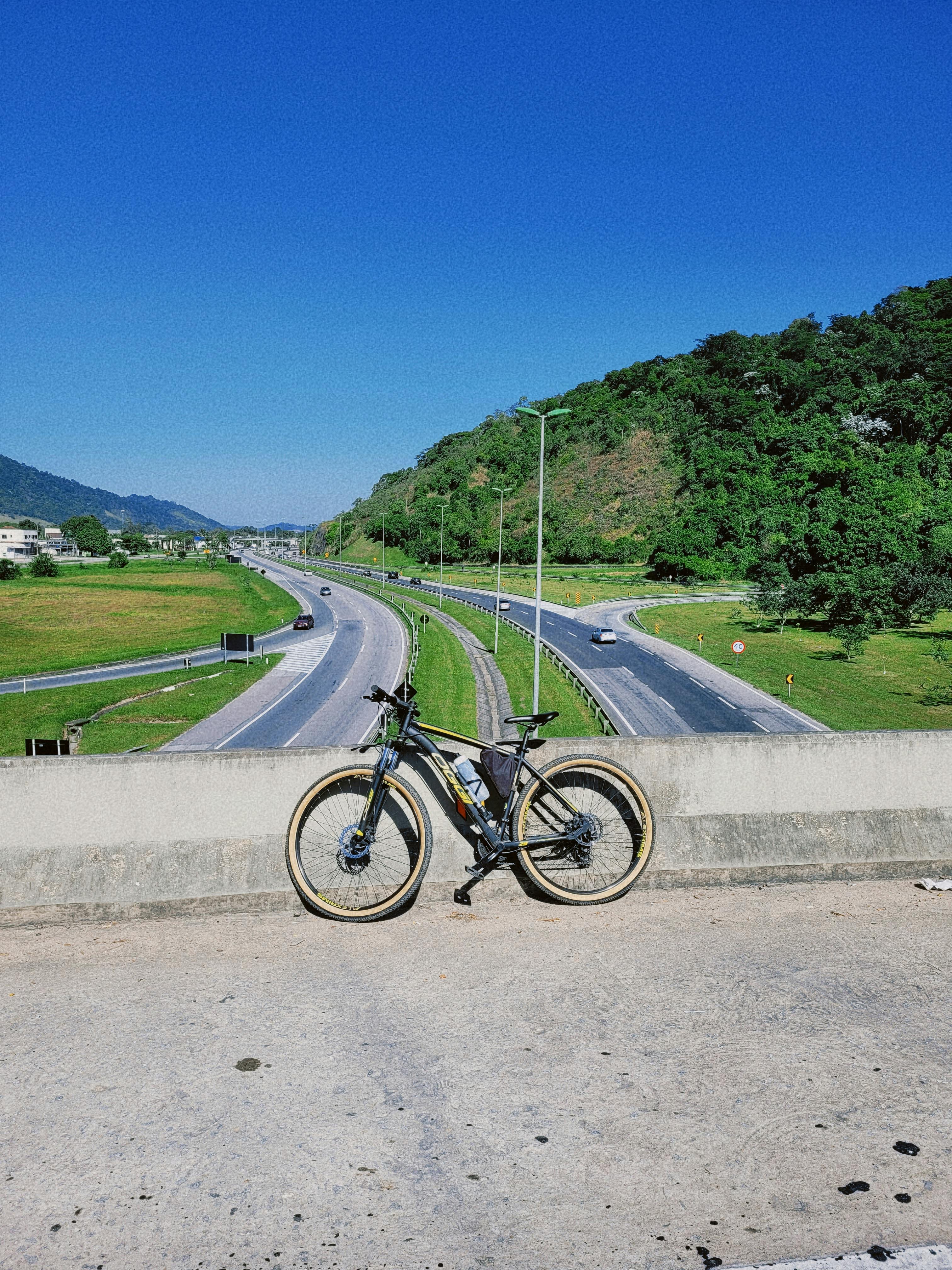 A bicycle stands on an overpass overlooking a scenic highway with green hills and a clear blue sky in the background.