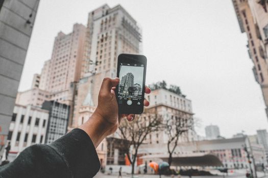 Crop anonymous person taking pictures of modern buildings located on urban street by using mobile phone during autumn cold day