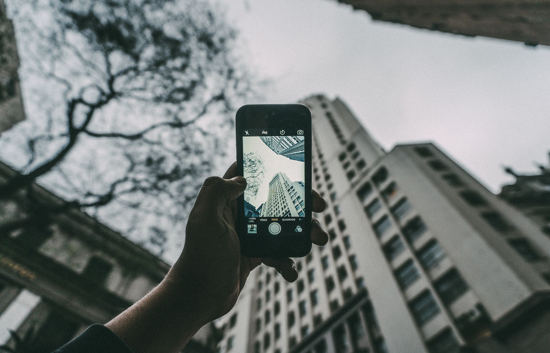 A hand holds a smartphone capturing city buildings from a low angle.