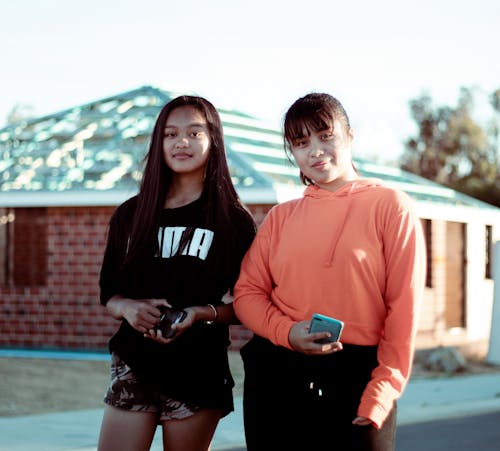 Two Women Wearing Orange Pullover Hoodie and Black T-shirt Standing in Front of Brown House