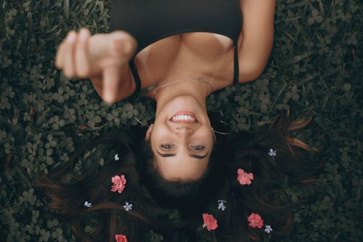 Smiling woman lying on grass with flowers in hair, pointing upwards.