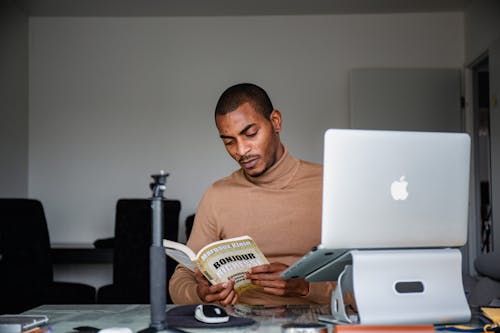 Man Sitting and Reading Book near Laptop