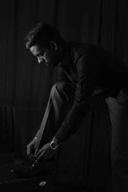 Фотография человека в классической рубашке и штанах, завязывающего шнурки в оттенках серого