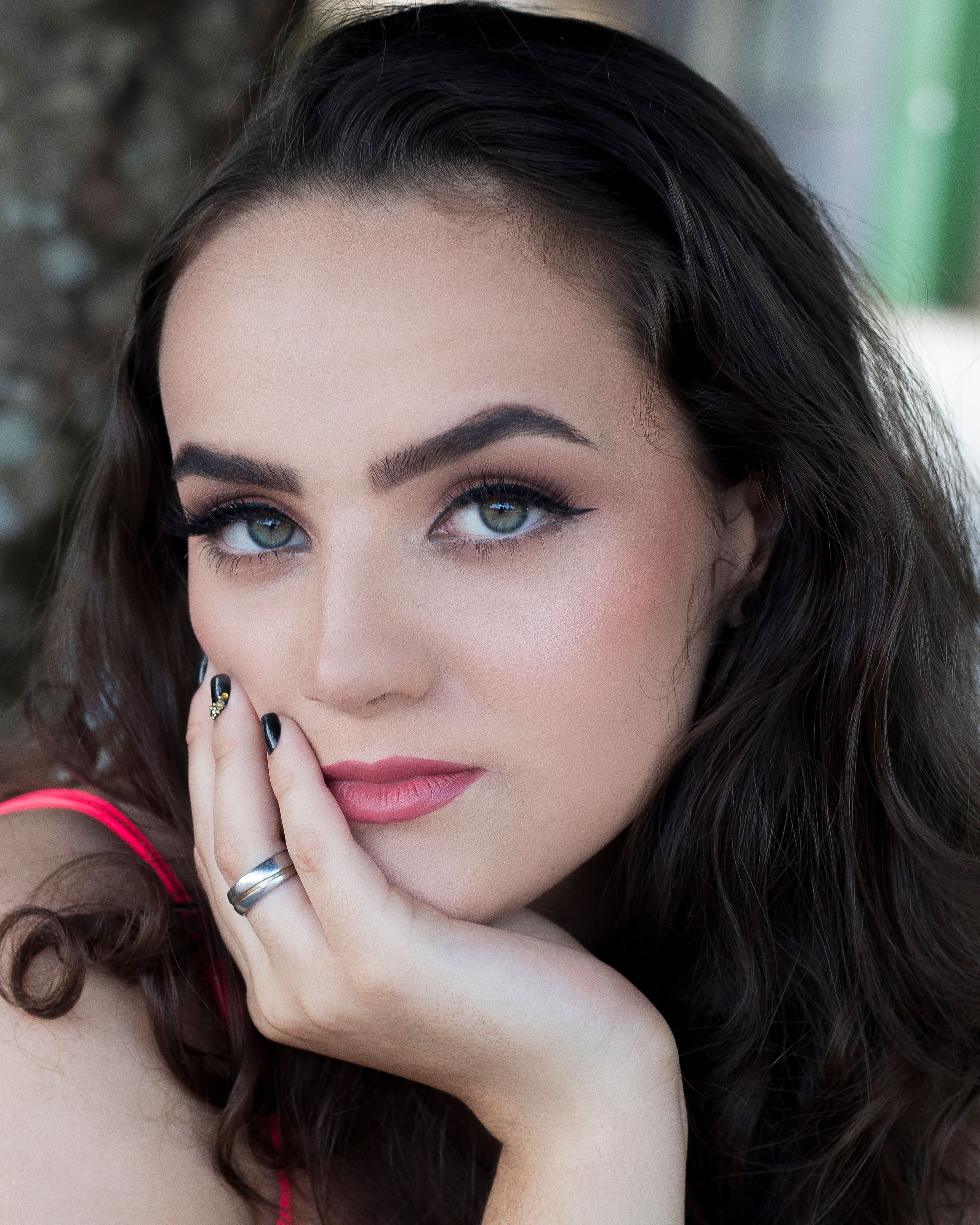 Close-up portrait of a brunette woman resting her chin on her hand with a thoughtful expression, showcasing makeup and jewelry.