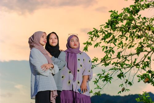 Free Three women in hijabs pose together under a colorful sunset sky, showcasing unity and fashion. Stock Photo