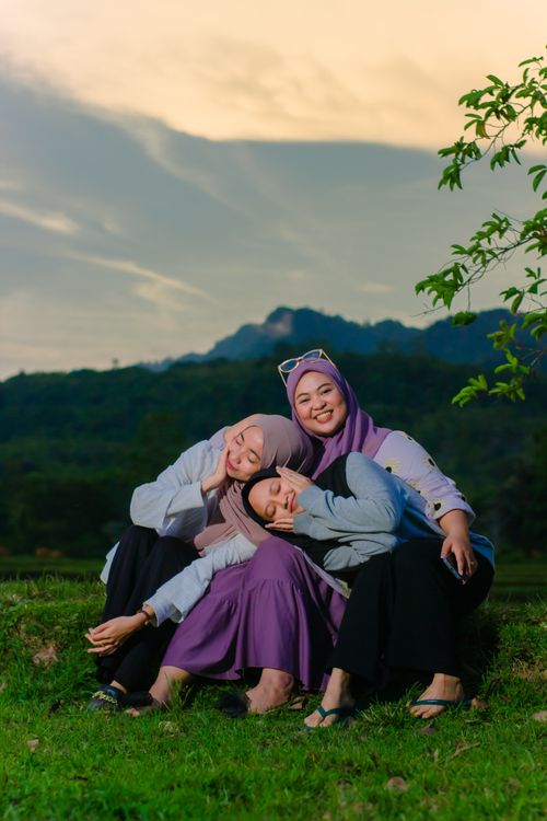 Free Three women wearing hijabs sitting on grass, smiling and relaxing together at dusk with a mountain backdrop. Stock Photo