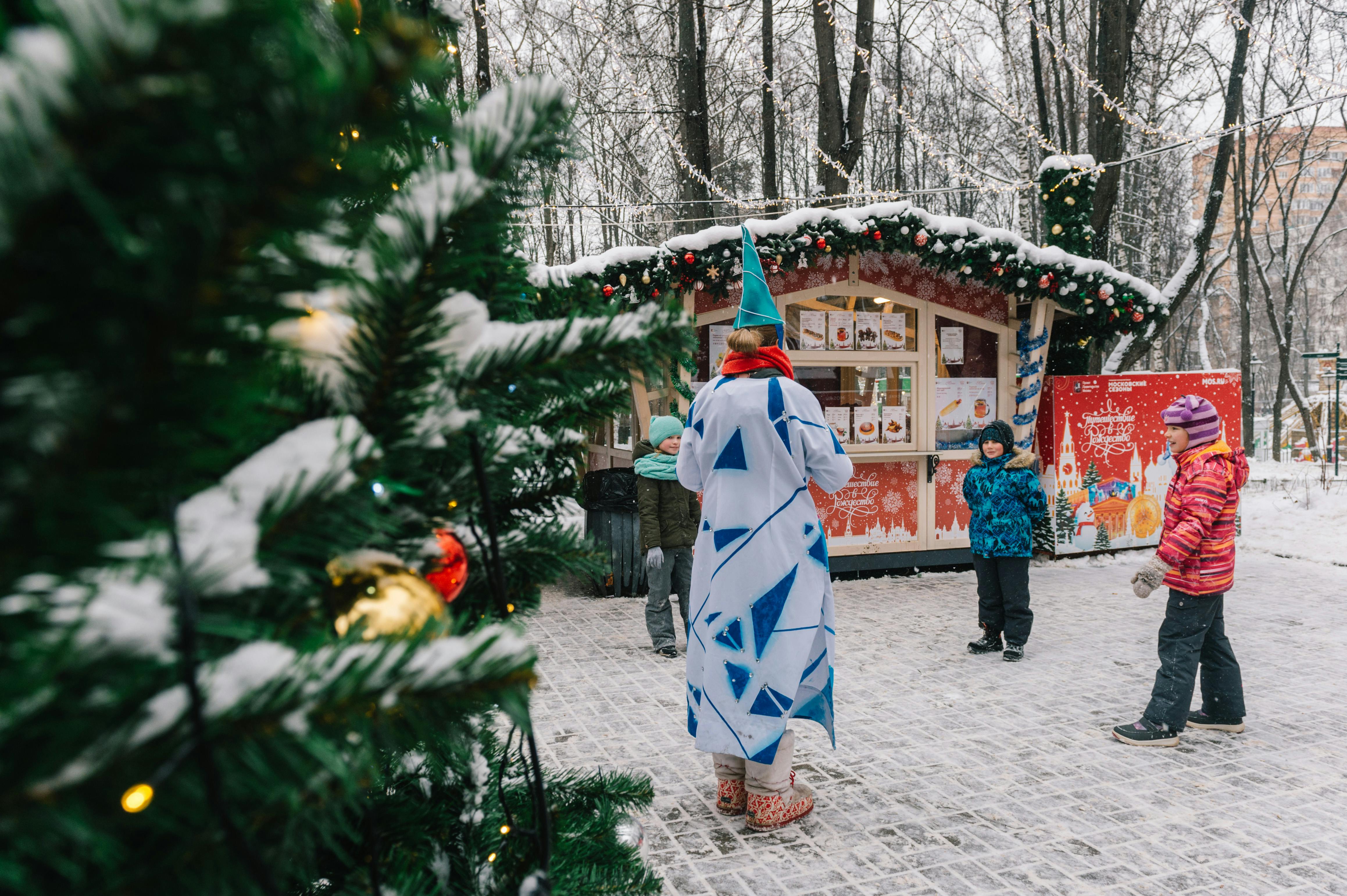 Children enjoying a snowy day at a festive Christmas market with decorations and a character in costume.