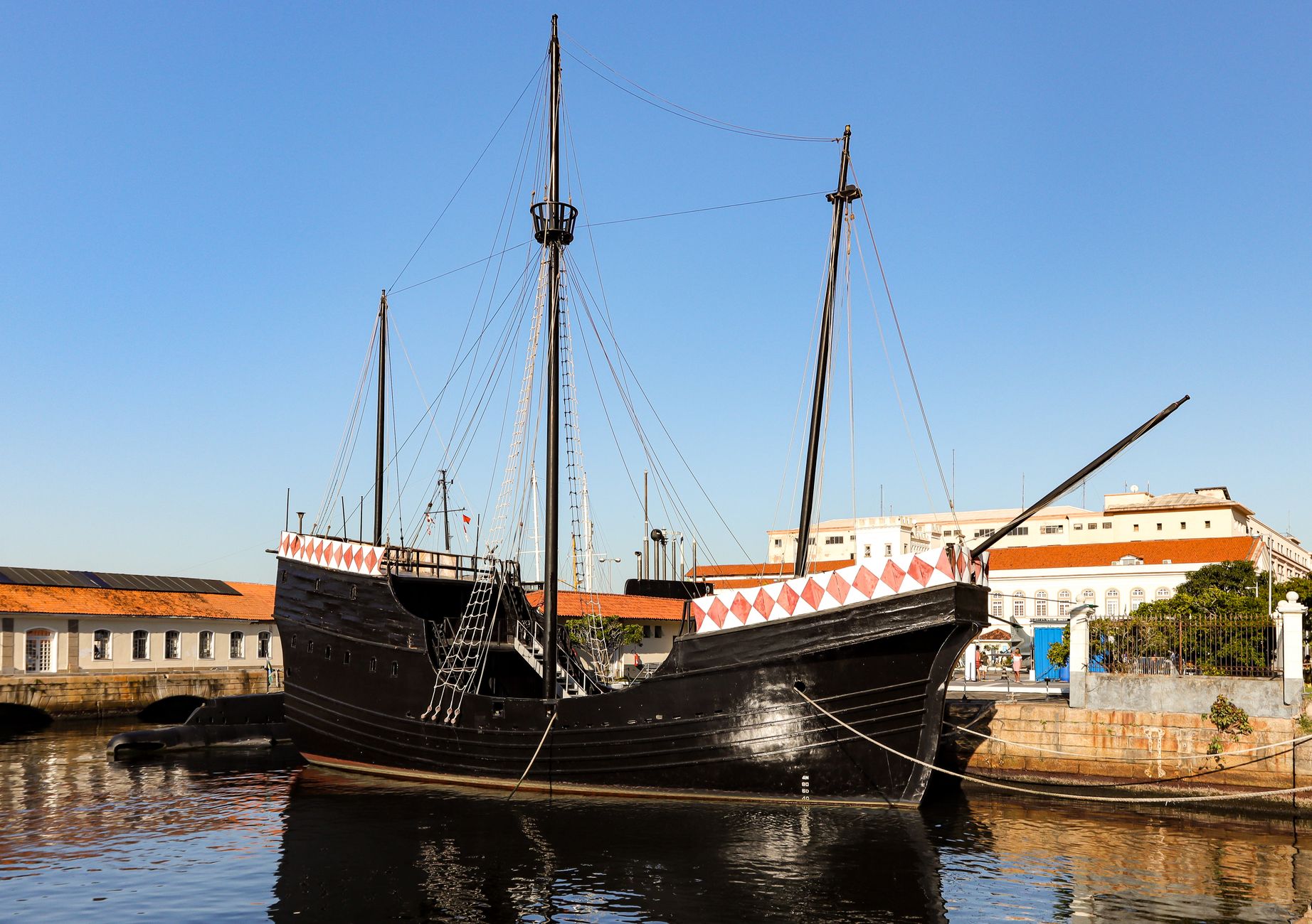 A historic black ship moored at a calm harbor, reflecting serene skies, captured in daylight.