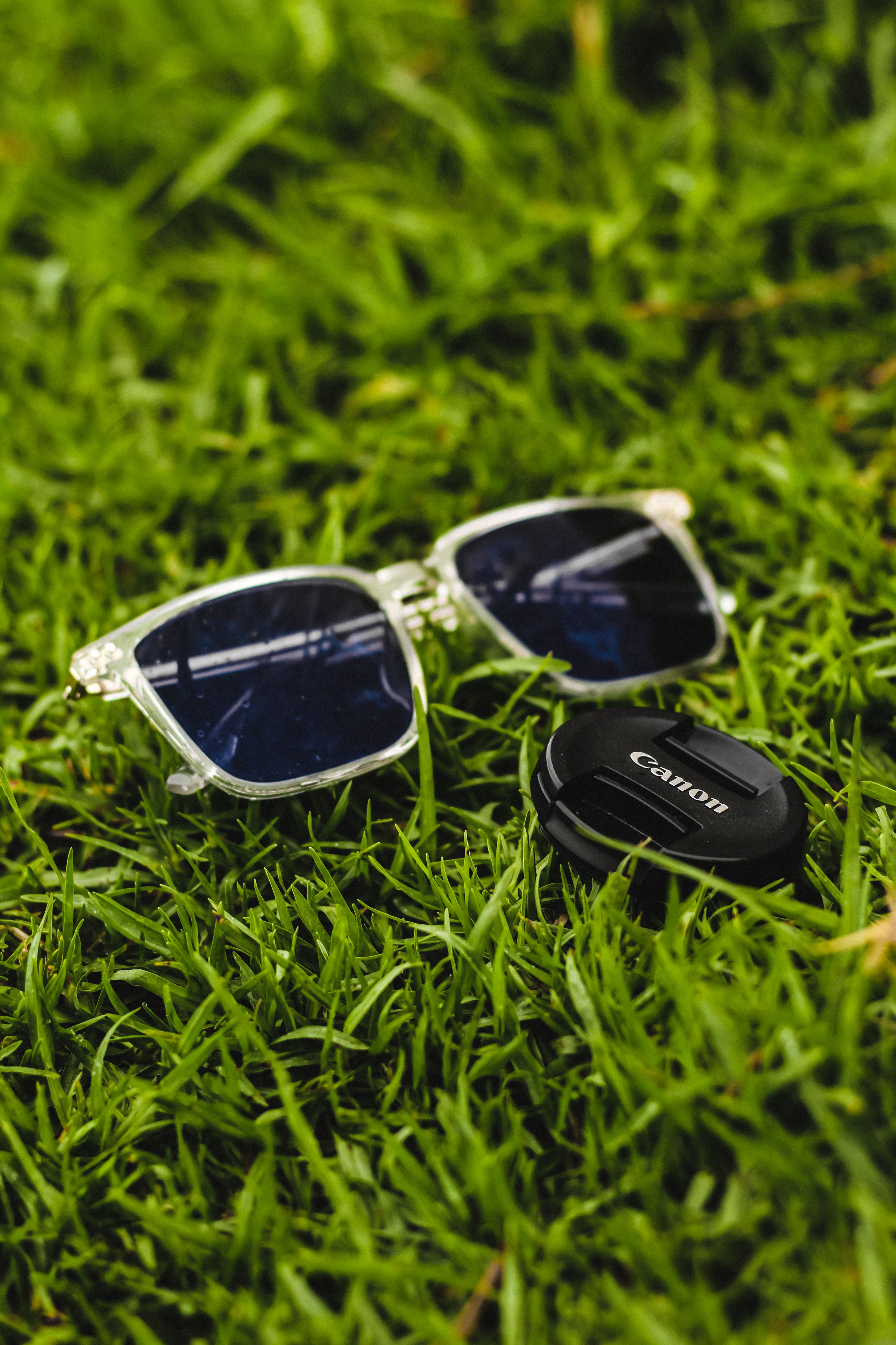 Close-up of sunglasses and Canon lens cap resting on vibrant green grass in Algeria.