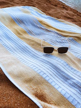 A beach towel with sunglasses lies on the sand by the shore. Ideal for summer themes.