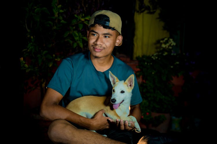 Young Man In Baseball Cap Holding A Funny Dog In His Lap