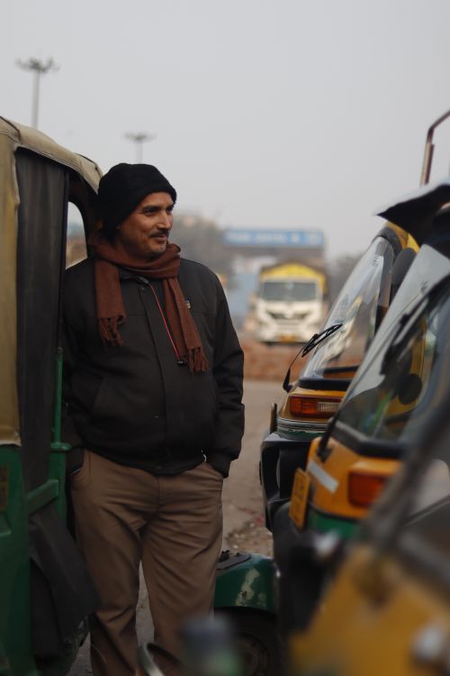 Free A man stands beside auto rickshaws in Delhi, India, wearing a scarf and jacket in the fog. Stock Photo