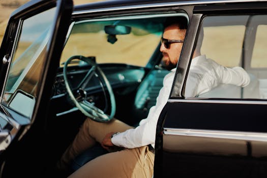 Stylish man in sunglasses sitting in a vintage car with the door open, outdoors in daylight.