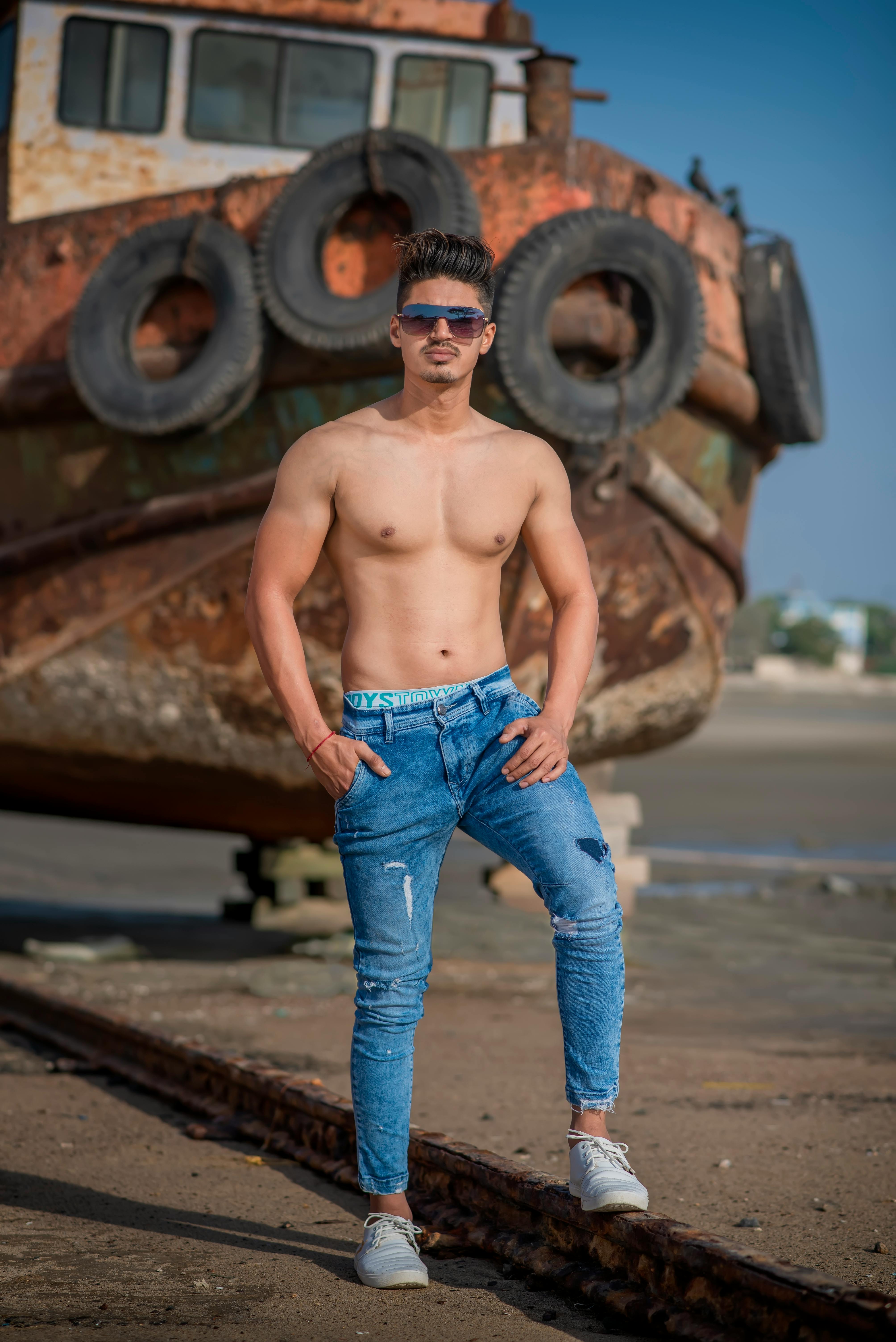 Topless man in jeans and sunglasses posing near a rusted ship on Mandvi Beach, India.