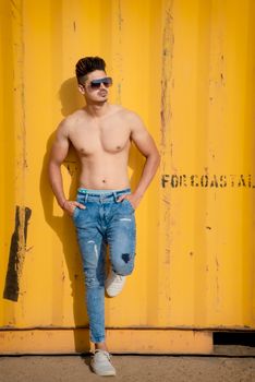 A young man models confidently shirtless, wearing jeans and sunglasses outdoors by a yellow container.