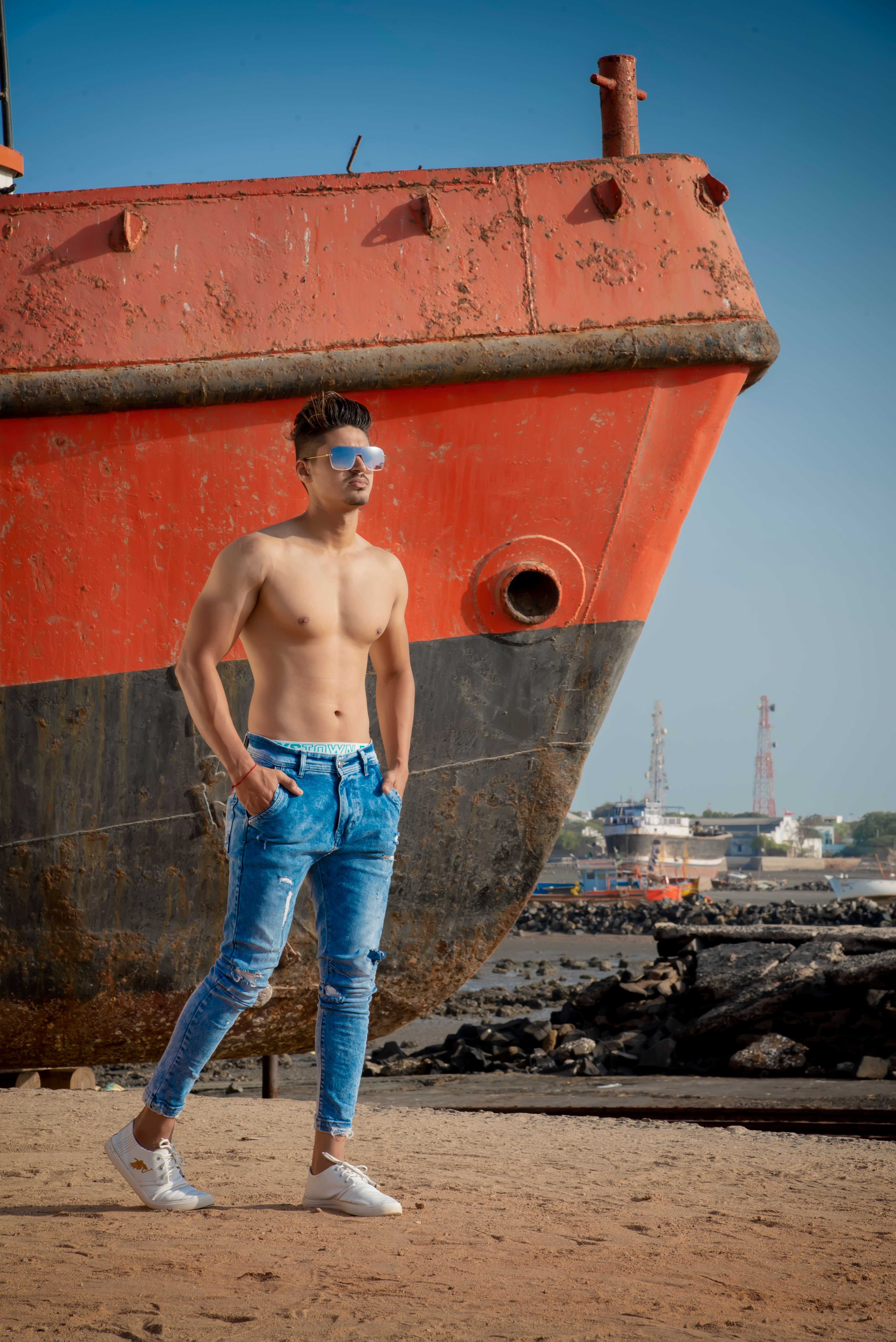 Stylish shirtless man in jeans and sunglasses poses by a ship near the beach in Mandvi, India.