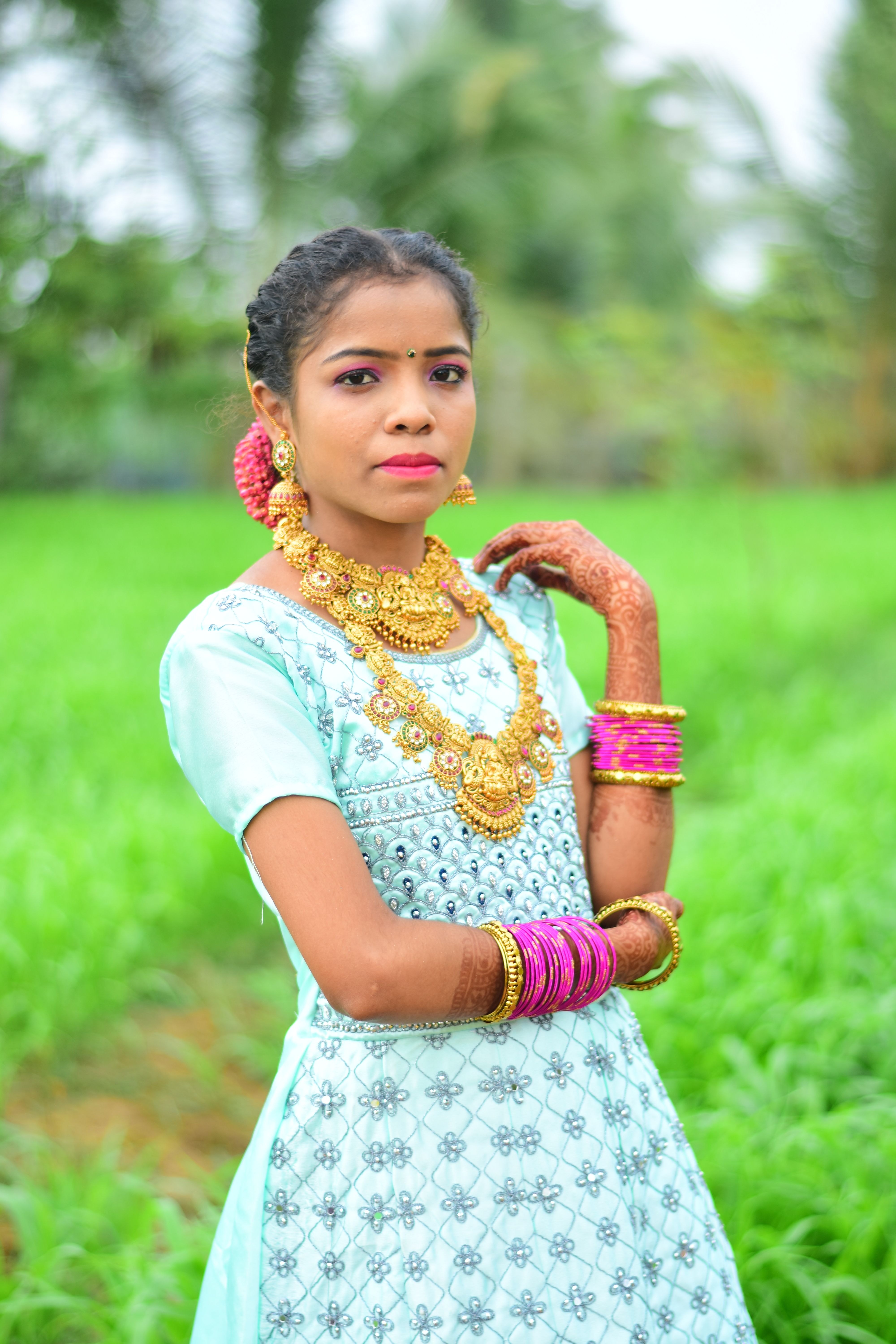 A South Asian woman stands gracefully in a field wearing traditional jewelry and attire, exuding elegance.