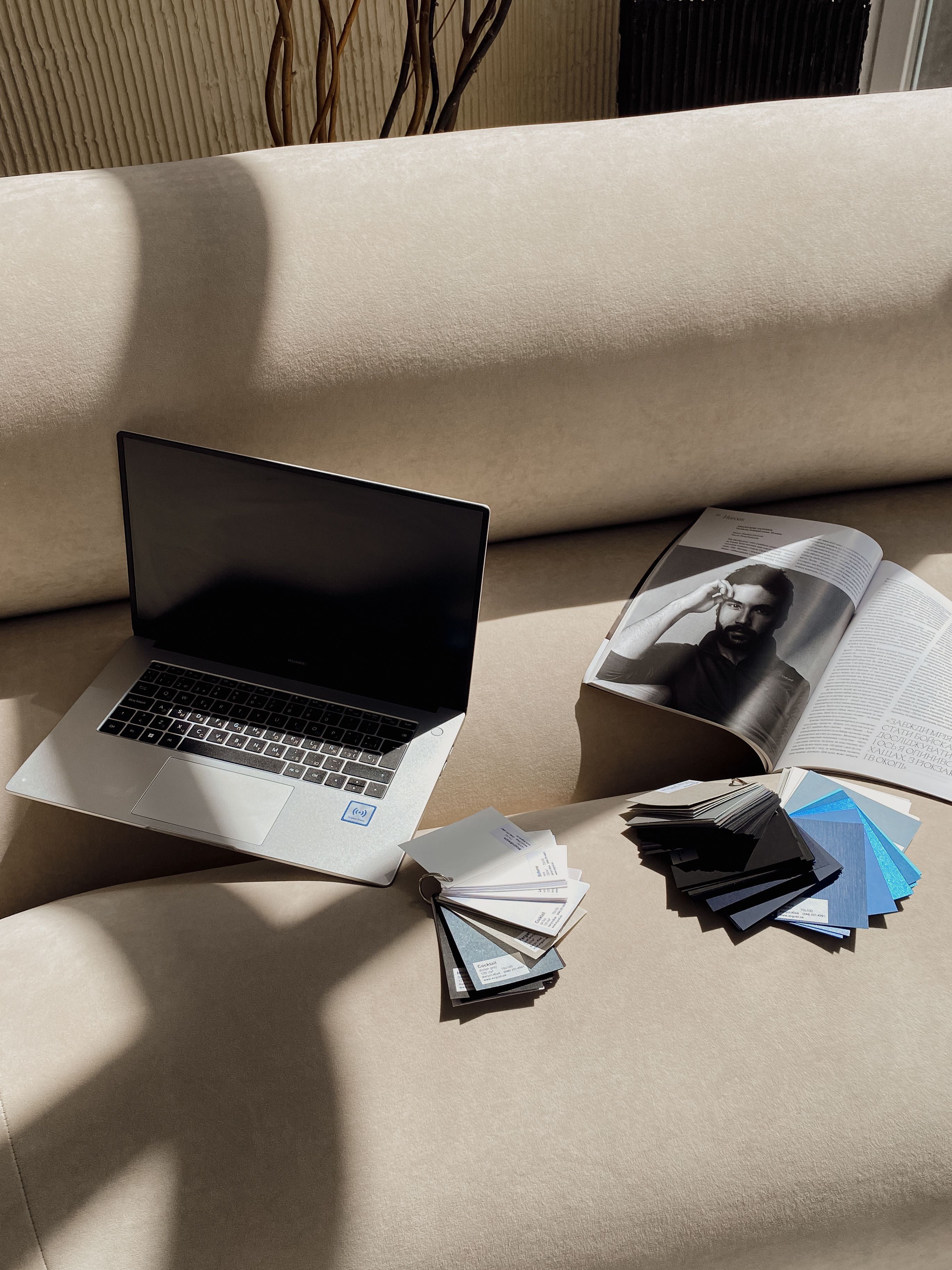 Modern workspace with laptop, magazine, and color swatches on a sofa in natural light.