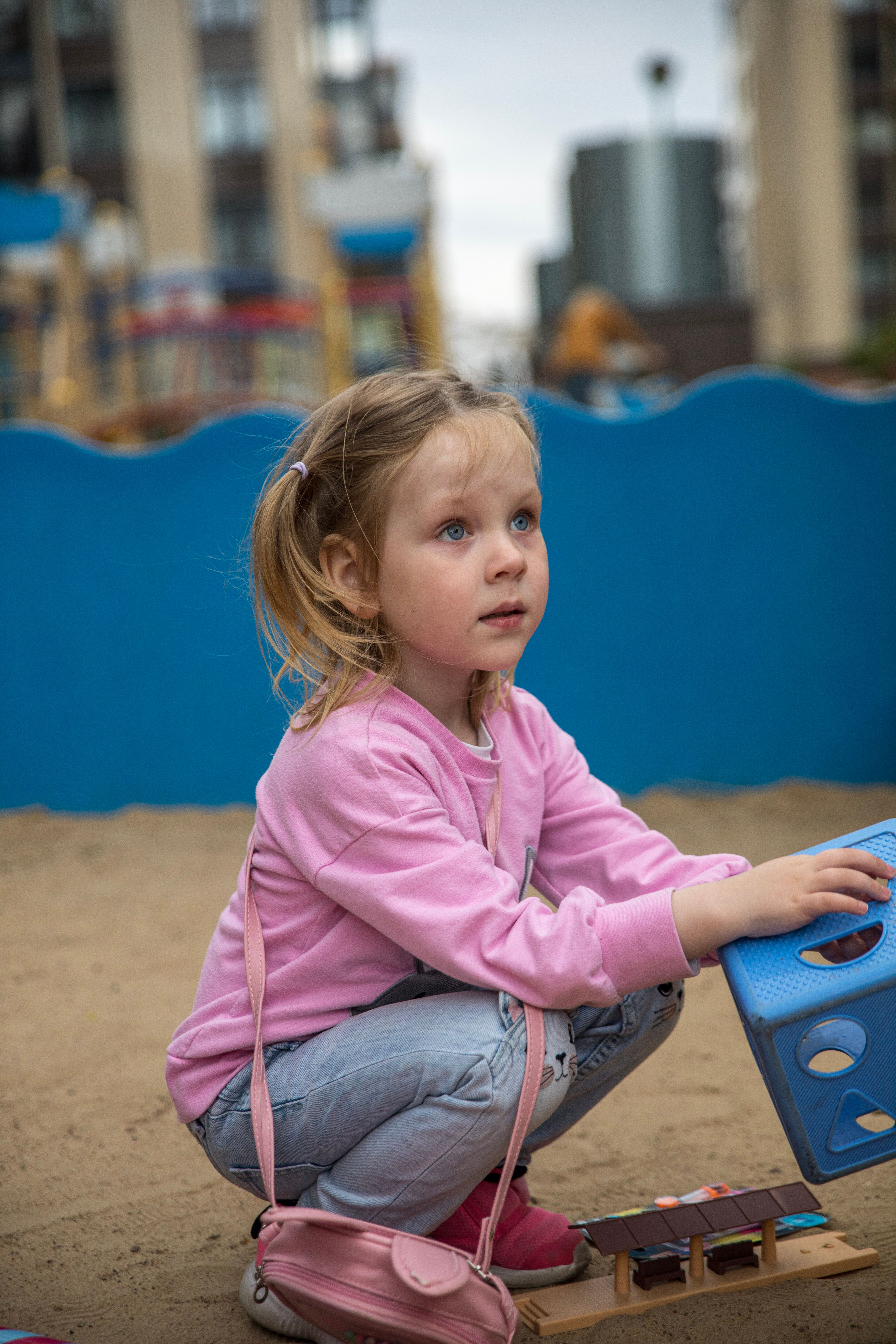 Blonde girl in pink squatting while playing in a city playground, surrounded by urban structures.