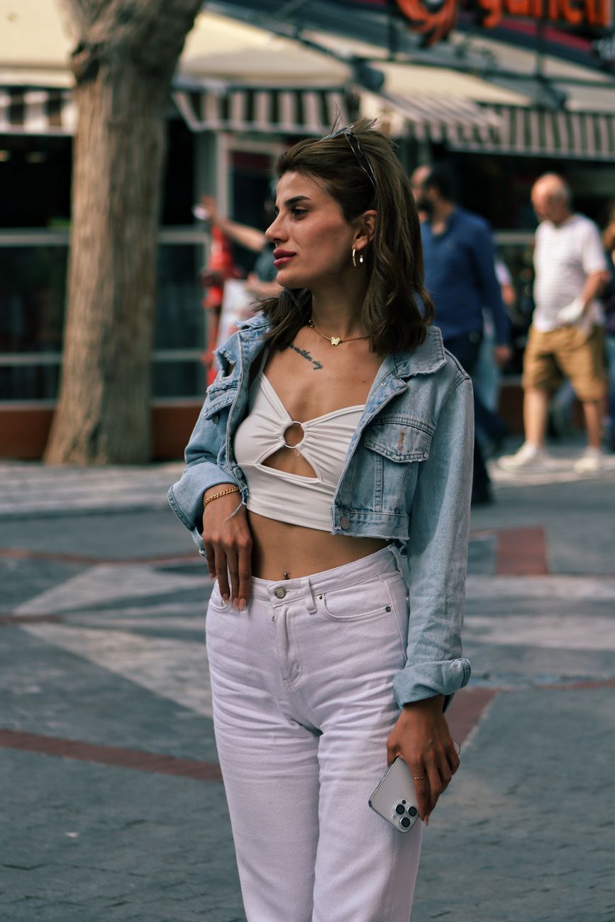 Mulher estilosa usa uma jaqueta jeans e segura um telefone enquanto está em pé ao ar livre em uma calçada da cidade.