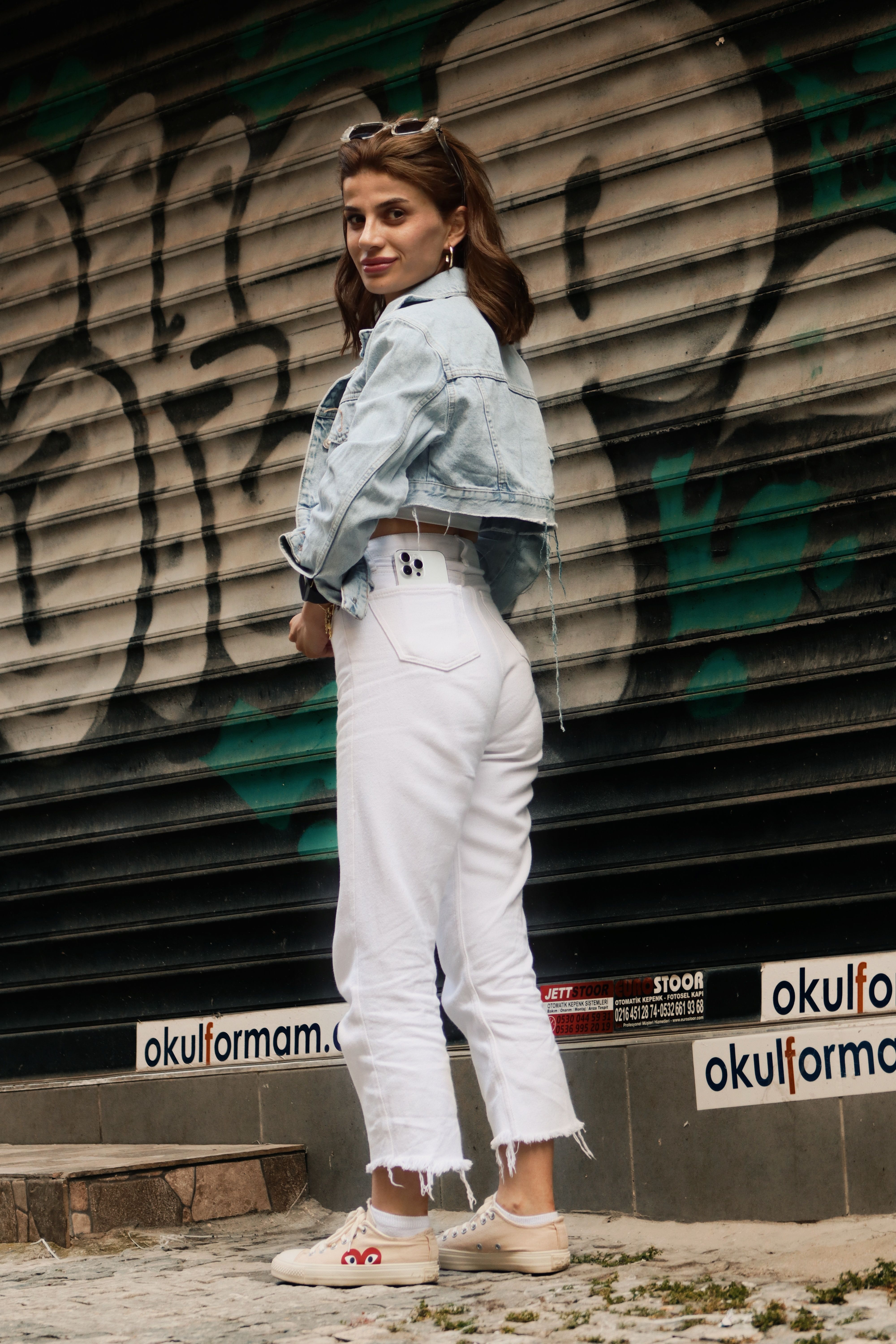 Young woman in jean jacket and white pants poses in front of a graffiti-covered garage door.
