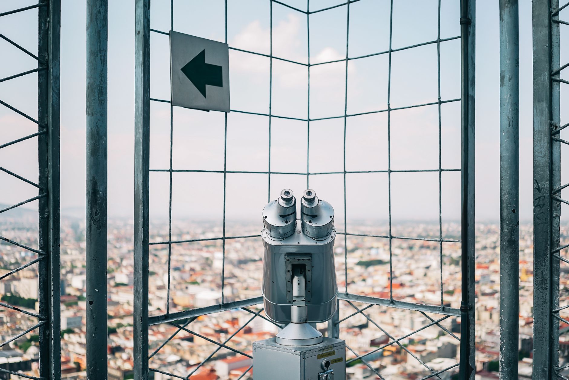 Binocular tower viewer overlooking a cityscape with a wire fence.