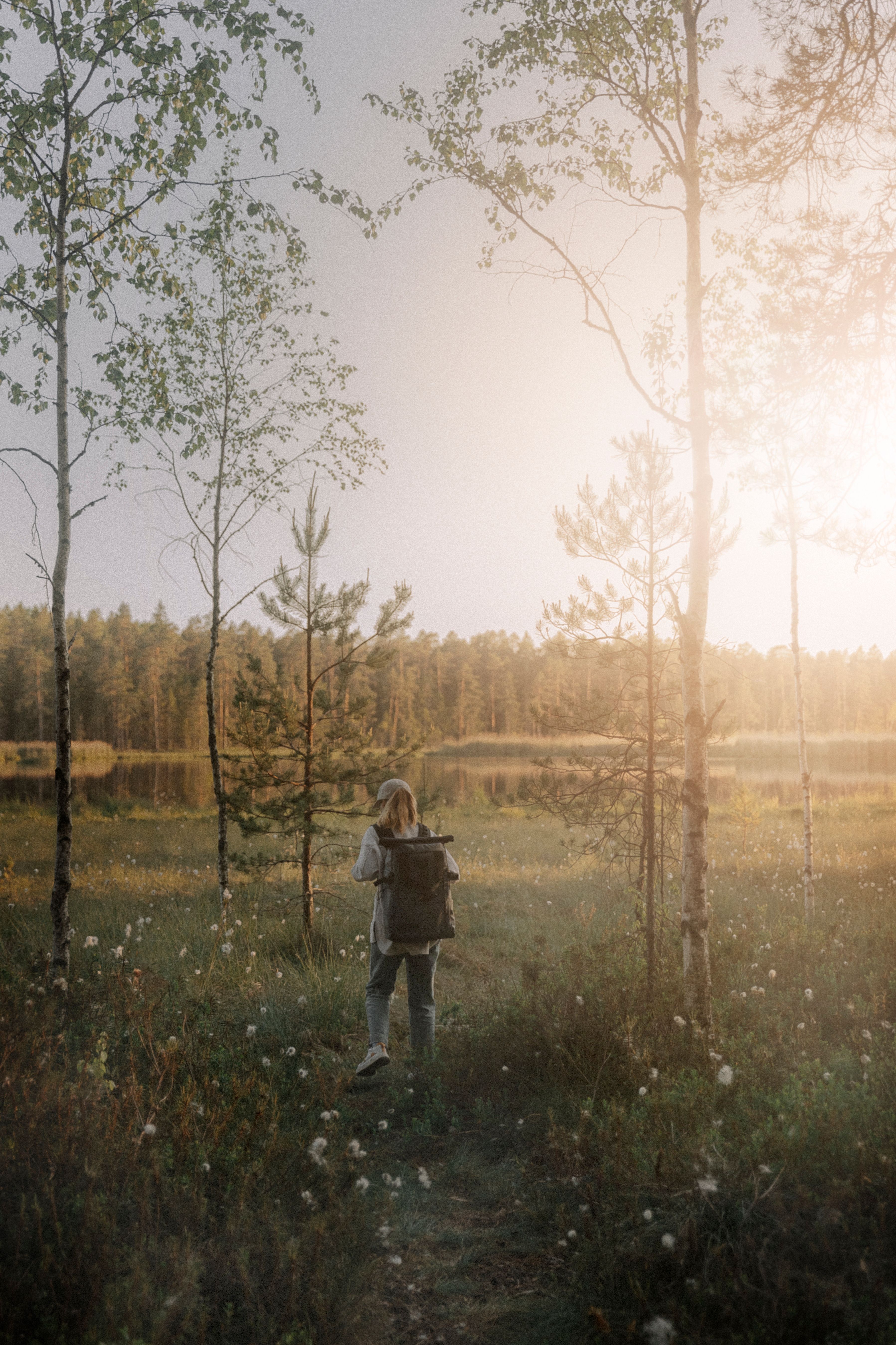 Person with a backpack standing by a calm forest lake in warm evening light