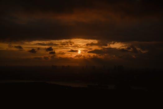 Dramatic orange sunset casting silhouettes over a city skyline with dark clouds.
