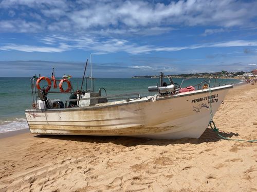 Free A weathered fishing boat beached on a sunlit sandy shore with calm sea and blue sky. Stock Photo