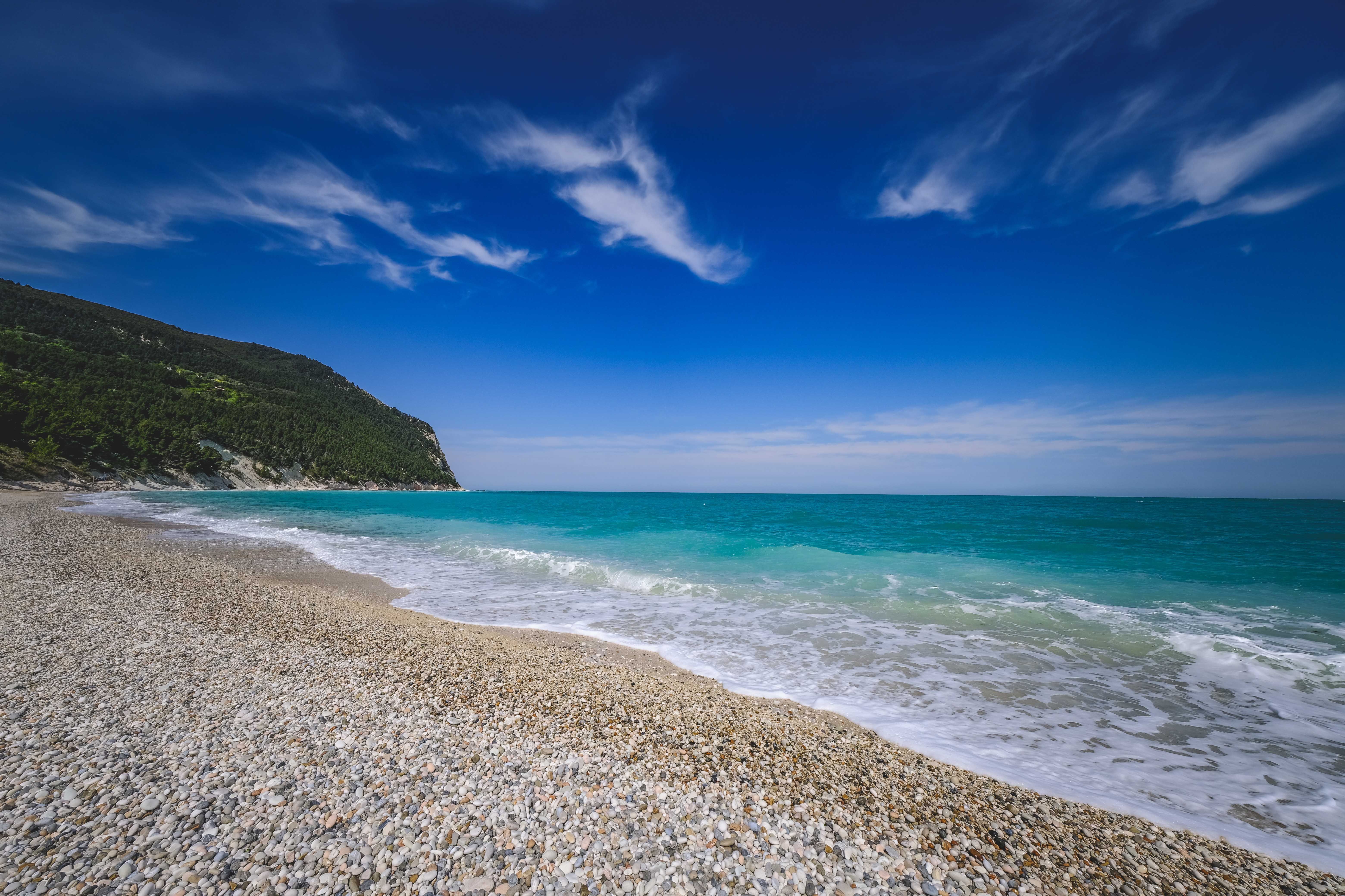 Pebble beach beside a turquoise sea, with a wooded headland beneath a bright blue sky.