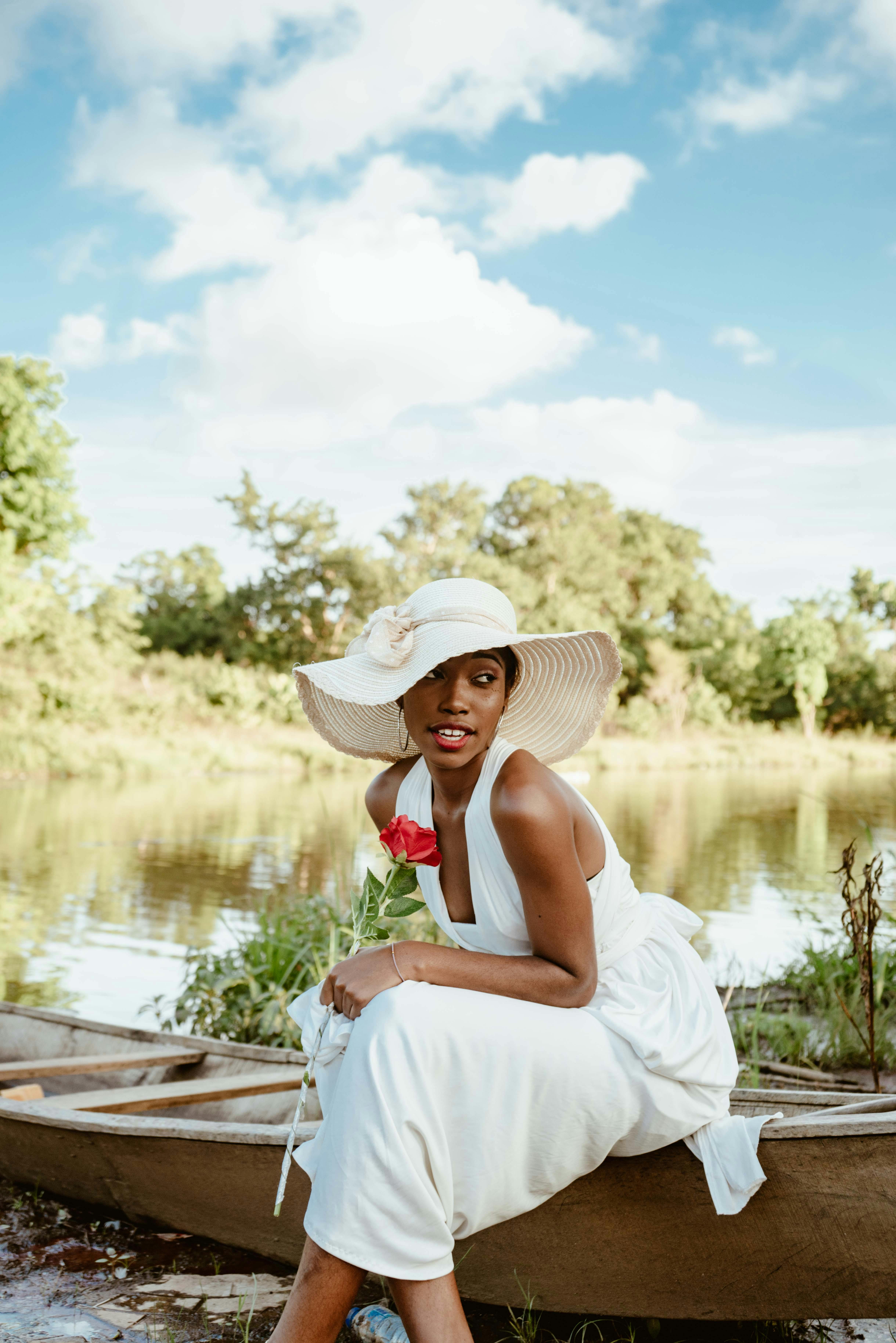 宁静的湖边，美丽的非洲妇女穿着白色连衣裙，戴着遮阳帽，手里拿着红玫瑰