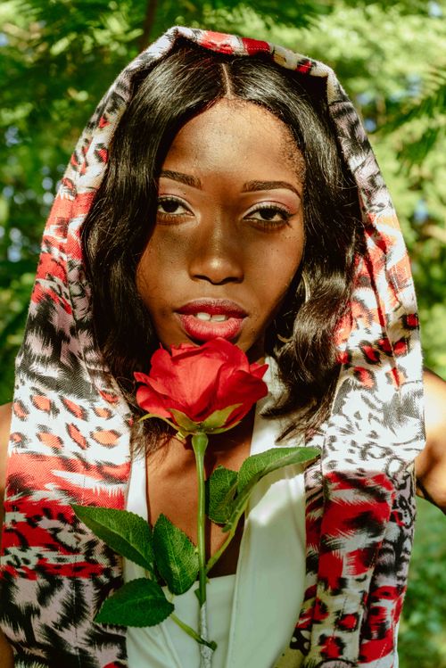 Free Stylish woman holds a red rose while wearing a vibrant scarf in a garden setting. Stock Photo