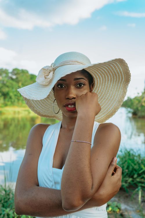 Free A fashionable woman in a wide-brimmed hat poses elegantly by a serene riverside with lush greenery. Stock Photo