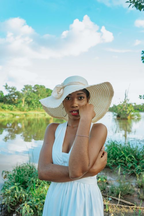 Free Stylish woman in white dress and hat posing by a scenic riverside under a bright summer sky. Stock Photo