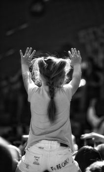 A young girl on shoulders enjoys a lively outdoor concert, arms raised in excitement.