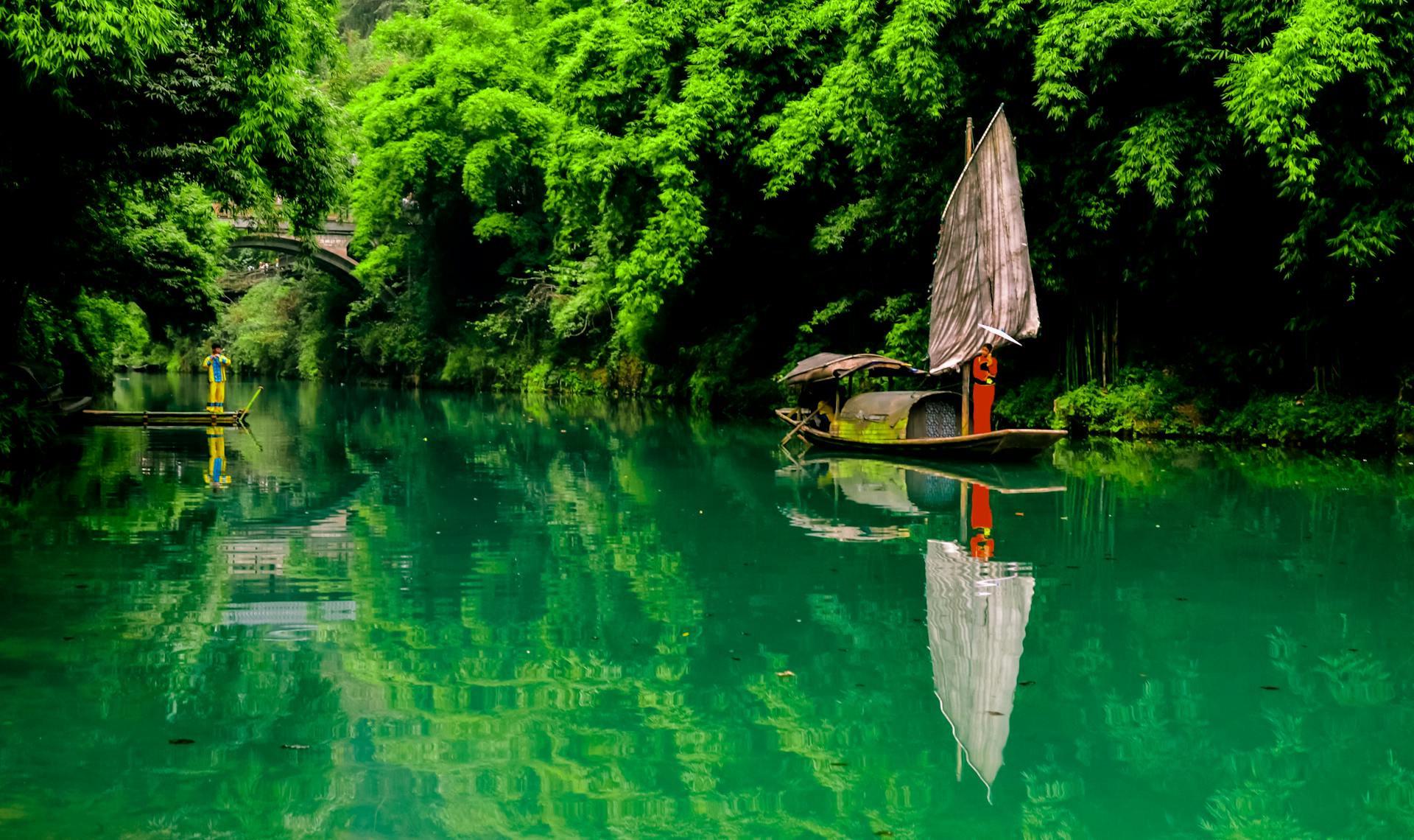 A traditional sailboat reflects on a calm river surrounded by lush green foliage.