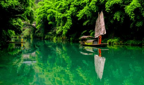 Free A traditional sailboat reflects on a calm river surrounded by lush green foliage. Stock Photo