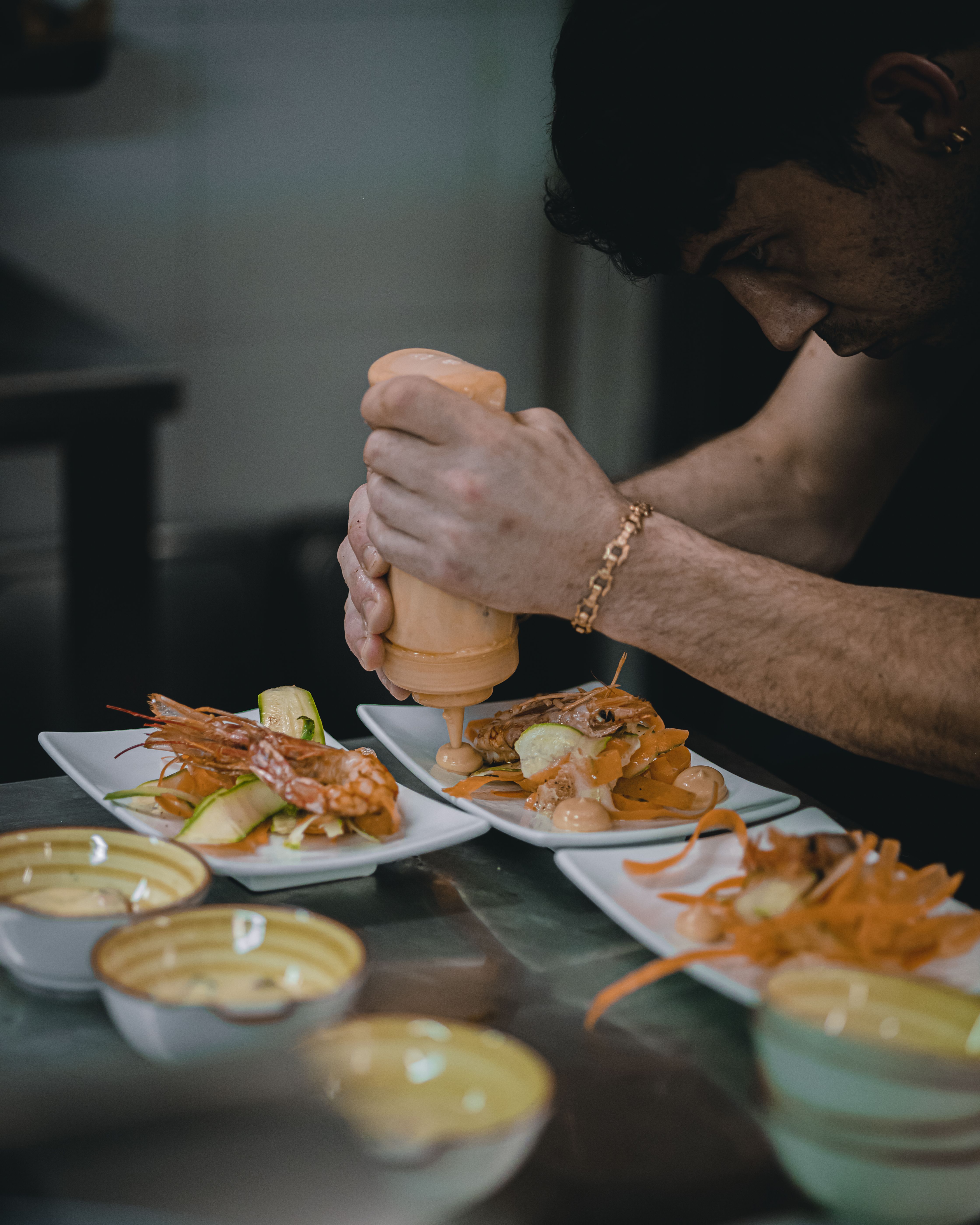 Professional chef preparing seafood, raw ingredients arranged precisely on a steel surface — shells, herbs, sea moss, and fermented paste.