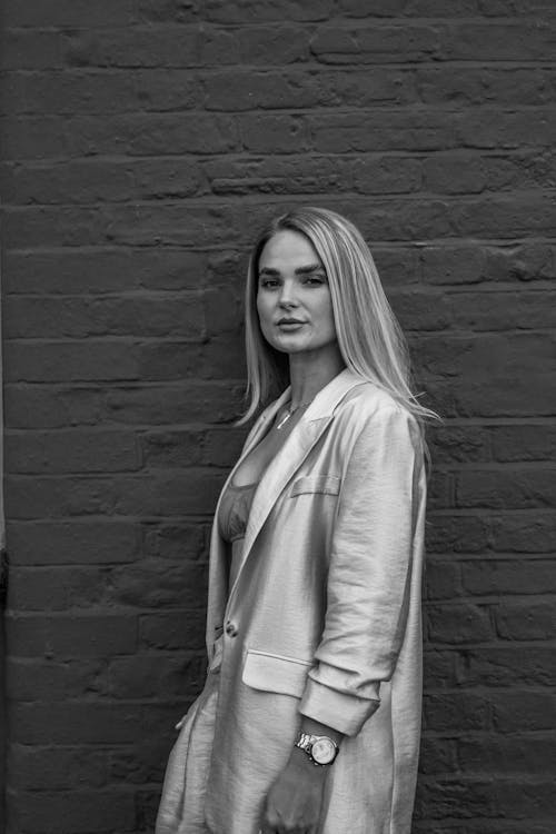 Black and White Photo of a Blonde Woman in Blazer and Palazzo Pants Standing by a Brick Wall