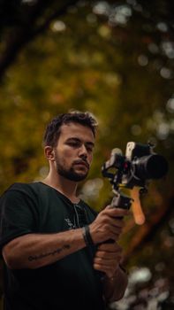 A young man in a t-shirt holding a camera on a tripod outdoors with blurred trees in the background.