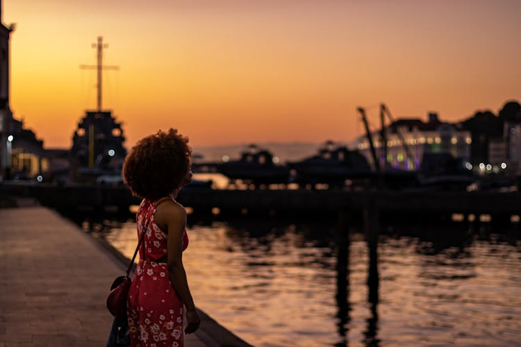 Young Woman In Red Flower Pattern Dress Enjoying Sunset Sky Over A Harbor