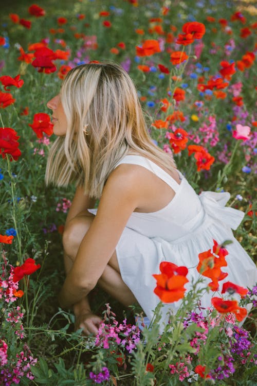 Blonde Woman in White Dress Squatting among Flowers on Meadow
