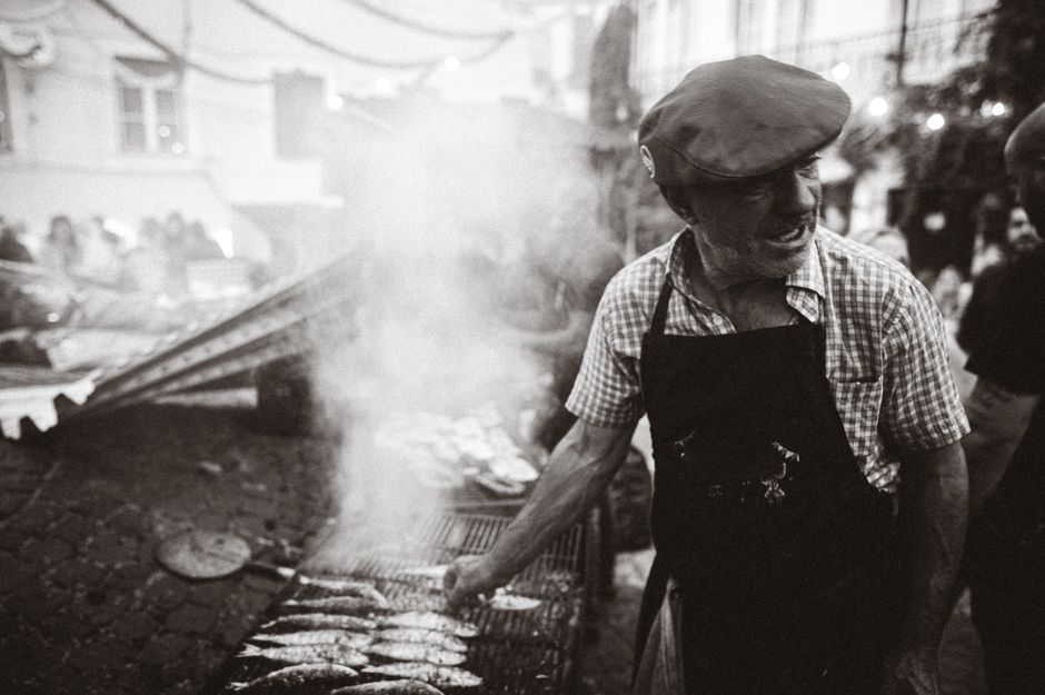 A street vendor grills fish in a bustling Lisbon market, surrounded by smoke and people.