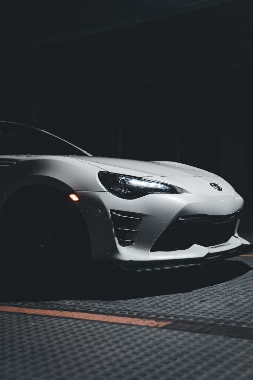 Free A stylish white sports car highlighted by headlights in a dimly lit parking lot scene. Stock Photo