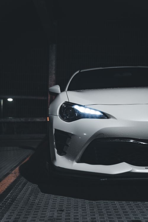 Free Front view of a white sports car with glowing headlights in a dimly lit parking lot at night. Stock Photo
